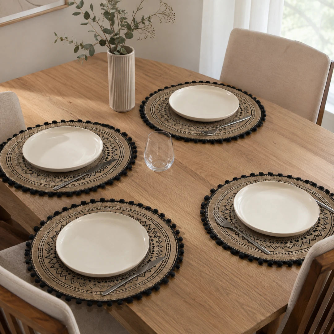 Josh & Tate - SunWeave™ Boho Mandala Placemat - Four round woven placemats with pom-pom trim and printed geometric patterns, each styled with a white plate, silver cutlery, and a clear glass on a wooden dining table in a bright room.