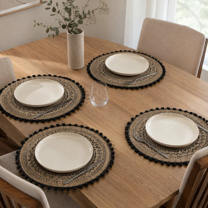 Josh & Tate - SunWeave™ Boho Mandala Placemat - Four round woven placemats with pom-pom trim and printed geometric patterns, each styled with a white plate, silver cutlery, and a clear glass on a wooden dining table in a bright room.