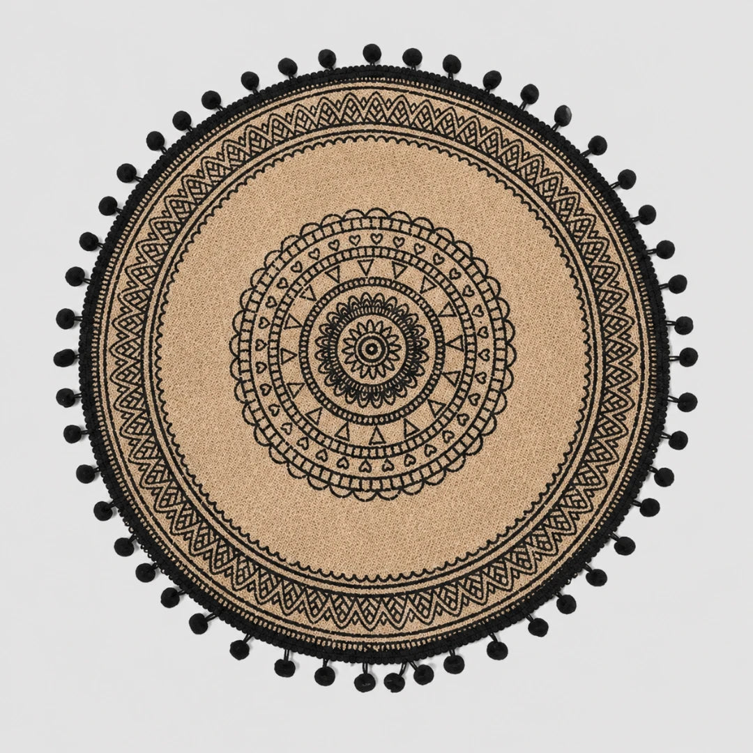 Josh & Tate - SunWeave™ Boho Mandala Placemat - A round beige woven placemat or rug with a black mandala pattern and black pom-pom trim, shown flat against a light gray background.