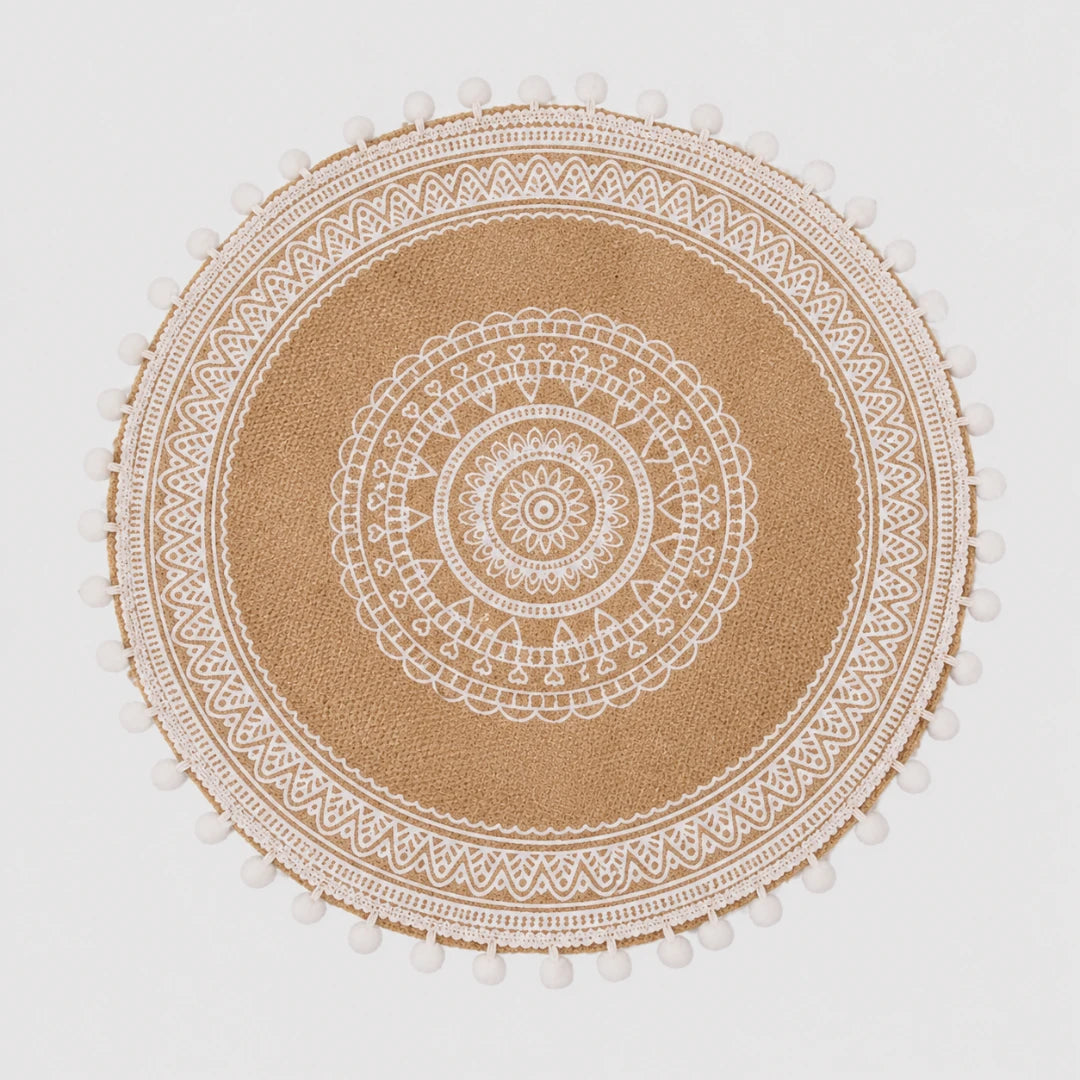 Josh & Tate - SunWeave™ Boho Mandala Placemat - A round natural jute placemat with a white printed mandala pattern and white pom-pom trim along the edge, shown flat against a light gray background.