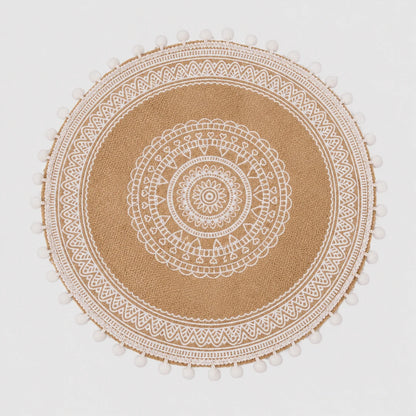 Josh & Tate - SunWeave™ Boho Mandala Placemat - A round natural jute placemat with a white printed mandala pattern and white pom-pom trim along the edge, shown flat against a light gray background.