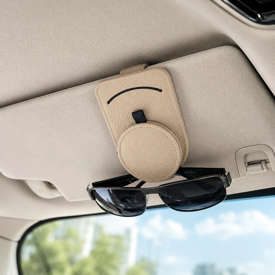 Josh & Tate - SunglassClip™ Car Visor Organizer - A beige sun visor holder with a smiley face design and a circular magnetic card slot, mounted on a car's sun visor with sunglasses resting underneath.