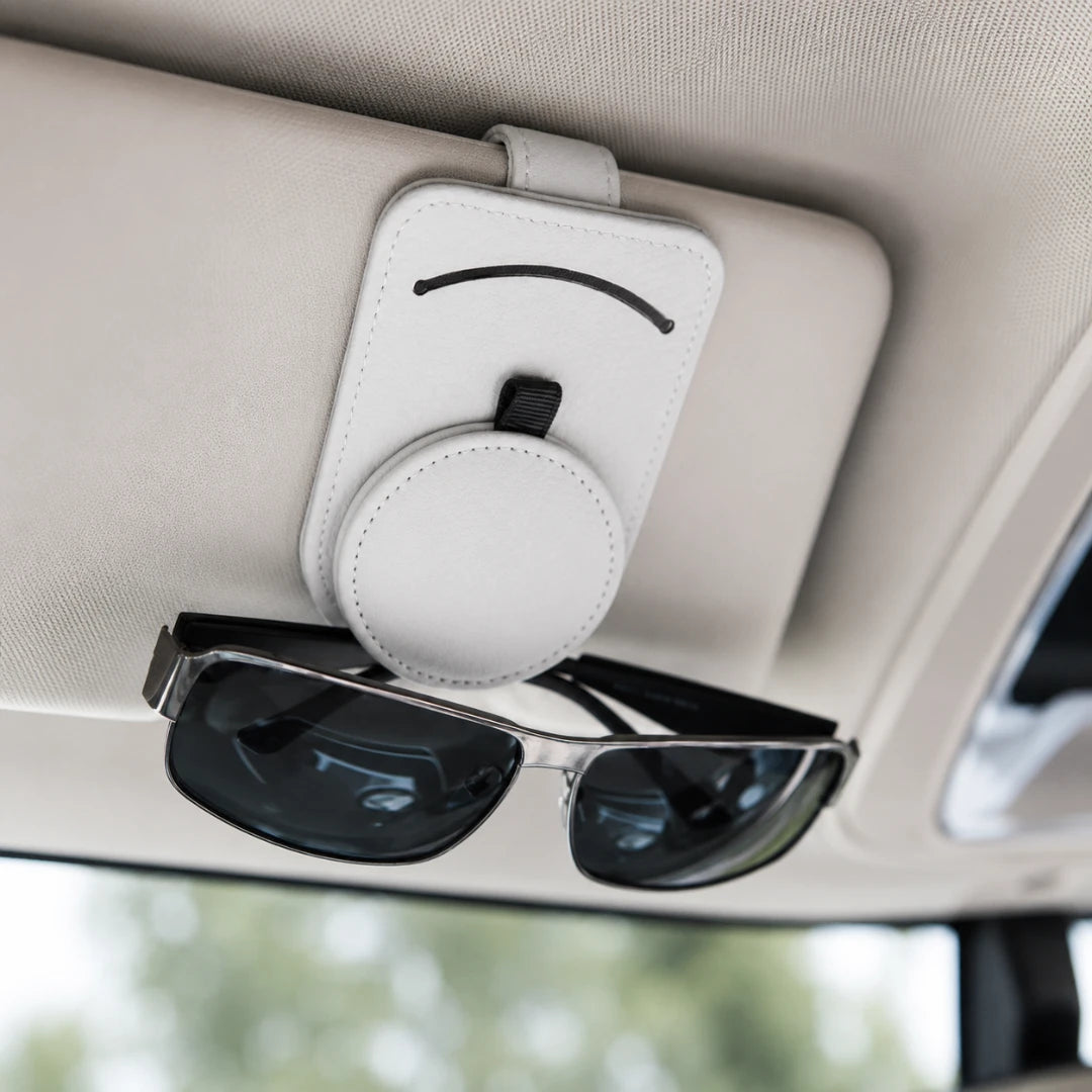 Josh & Tate - SunglassClip™ Car Visor Organizer - A white circular magnetic sunglasses holder attached to a beige car sun visor, holding a pair of black-framed sunglasses.