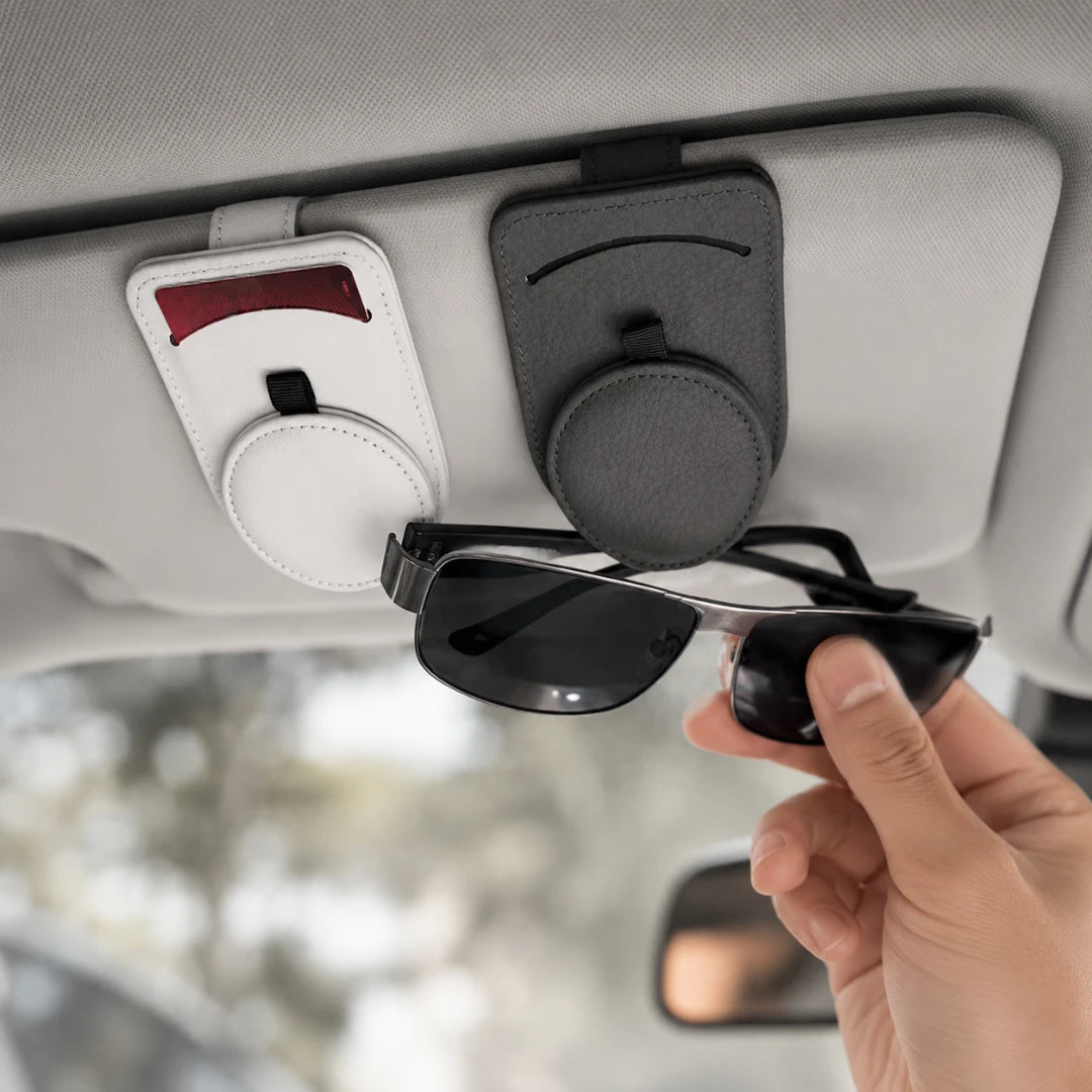 Josh & Tate - SunglassClip™ Car Visor Organizer - A hand holding a pair of black sunglasses under a light gray car sun visor, showing two magnetic visor-mounted holder cases in white and dark gray. One holder contains a red card, and the other holds the sunglasses.