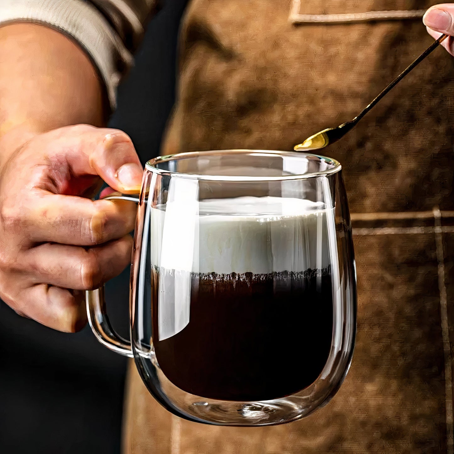 Person holding a glass mug with coffee and using a spoon to add a substance, possibly cream or sugar.