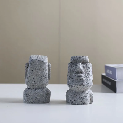 Two stone-like sculptures resembling Moai on a white surface with a neutral background