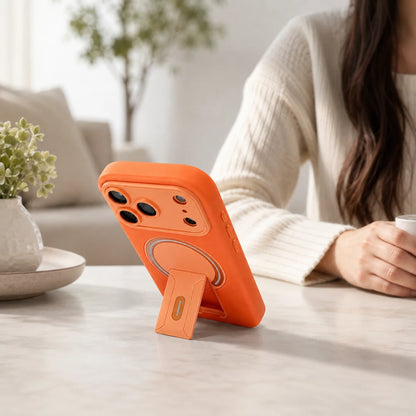 Josh & Tate - TiltPod™ iPhone Case - An orange phone case with an integrated kickstand stands on a light marble table in a bright, modern living room. A woman wearing a cream ribbed sweater is seated in the background, holding a cup.