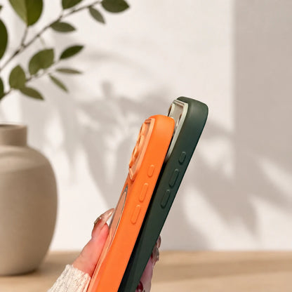 Josh & Tate - TiltPod™ iPhone Case - A hand holds two slim smartphone cases, one bright orange and one dark green, against a softly lit indoor background with a vase and leafy branch.