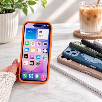 Josh & Tate - TiltPod™ iPhone Case - A hand holds an iPhone with a bright orange protective case on a white marble tabletop. In the background, other phone cases in blue, green, and beige are stacked on a white notebook next to an iced coffee and a potted plant.