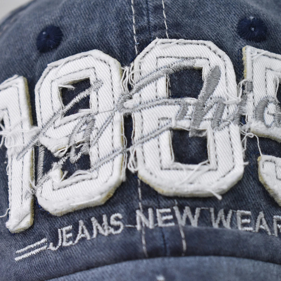 Josh & Tate - TrailFade™ '85 Cap - Close-up of a distressed denim baseball cap featuring large white embroidered '100' letters with frayed details, a gray signature overlay, and 'JEANS NEW WEAR' embroidered below.