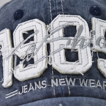Josh & Tate - TrailFade™ '85 Cap - Close-up of a distressed denim baseball cap featuring large white embroidered '100' letters with frayed details, a gray signature overlay, and 'JEANS NEW WEAR' embroidered below.