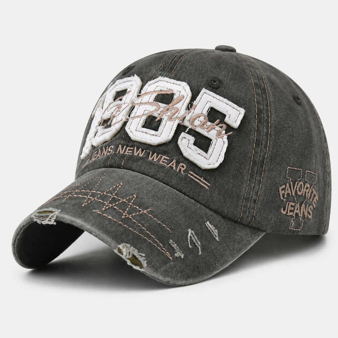 Josh & Tate - TrailFade™ '85 Cap - A distressed dark gray washed denim baseball cap featuring '1985' embroidered on the front, 'FABRIC' in script, and 'JEANS NEW WEAR' text, with 'FAVORITE JEANS' embroidered on the side.
