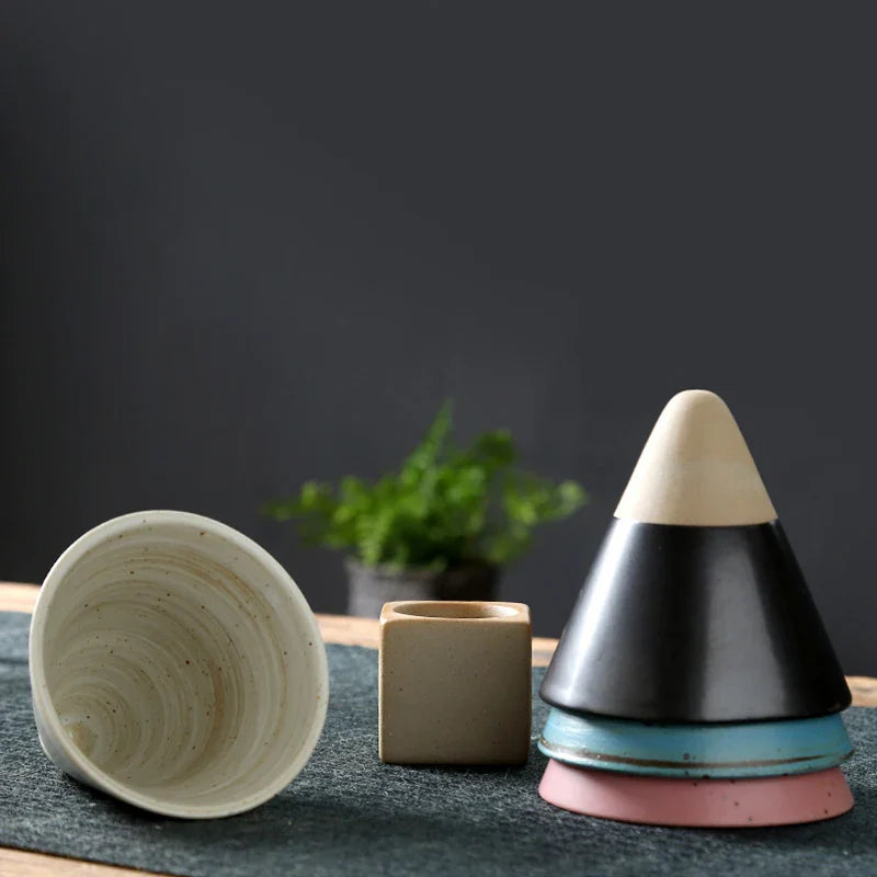 Set of ceramic items including a cone, cup, and cube on a dark surface with a plant in the background.