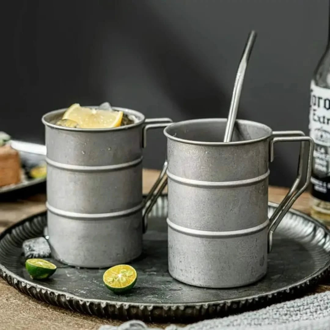 Josh & Tate - Vintage Steel Mug (13oz / 380mL) - Two metal mugs with handles on a dark tray, containing lemon slices and a bottle of liquor in the background.