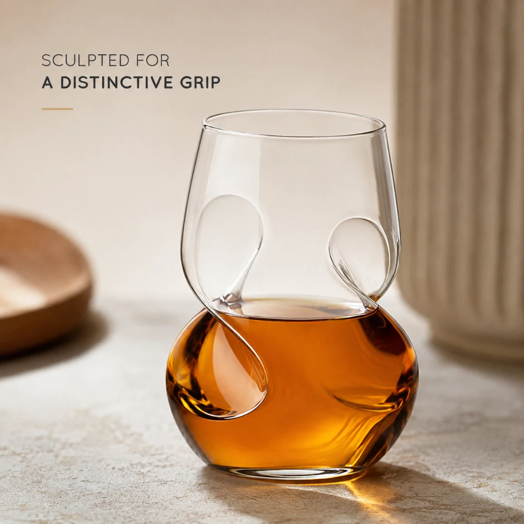 Josh & Tate - VortexPour™ Spiral Wine Glass - A sculpted clear glass tumbler with organic looped details, partially filled with amber liquid, set on a stone surface with warm neutral background elements and the text 'SCULPTED FOR A DISTINTIVE GRIP'.