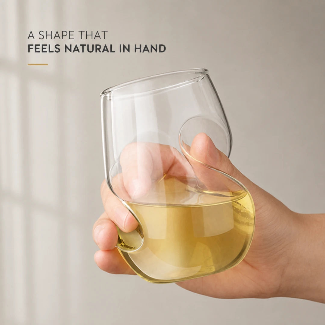 Josh & Tate - VortexPour™ Spiral Wine Glass - A hand holding a clear stemless wine glass with a unique wavy, integrated handle design, partially filled with pale yellow liquid. The background is a soft beige wall with text in the upper left reading 'A SHAPE THAT FEELS NATURAL IN HAND'.