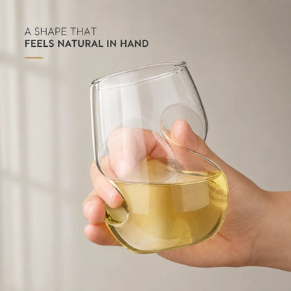 Josh & Tate - VortexPour™ Spiral Wine Glass - A hand holding a clear stemless wine glass with a unique wavy, integrated handle design, partially filled with pale yellow liquid. The background is a soft beige wall with text in the upper left reading 'A SHAPE THAT FEELS NATURAL IN HAND'.