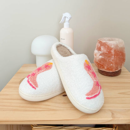 Pair of white slippers with pink sole on a wooden surface, surrounded by a bottle, mushroom-shaped object, and salt lamp.