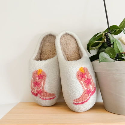 Pair of white slippers with pink cowboy boot design on a wooden surface with a plant in the background.