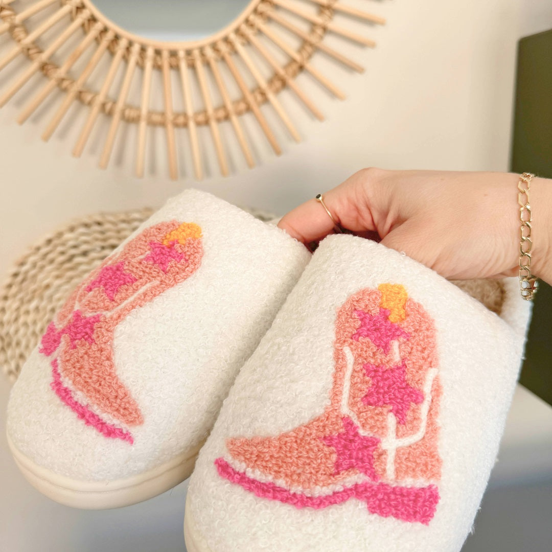 White slippers with pink cowboy boot design held by a hand against a neutral background