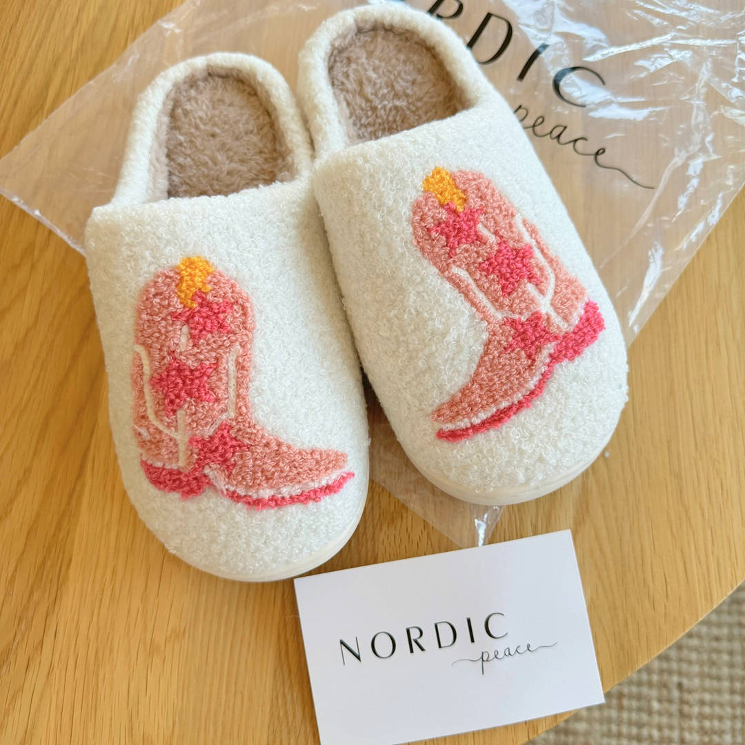 Pair of white slippers with pink boot design on a wooden surface, featuring the brand 'Nordic Peace'.