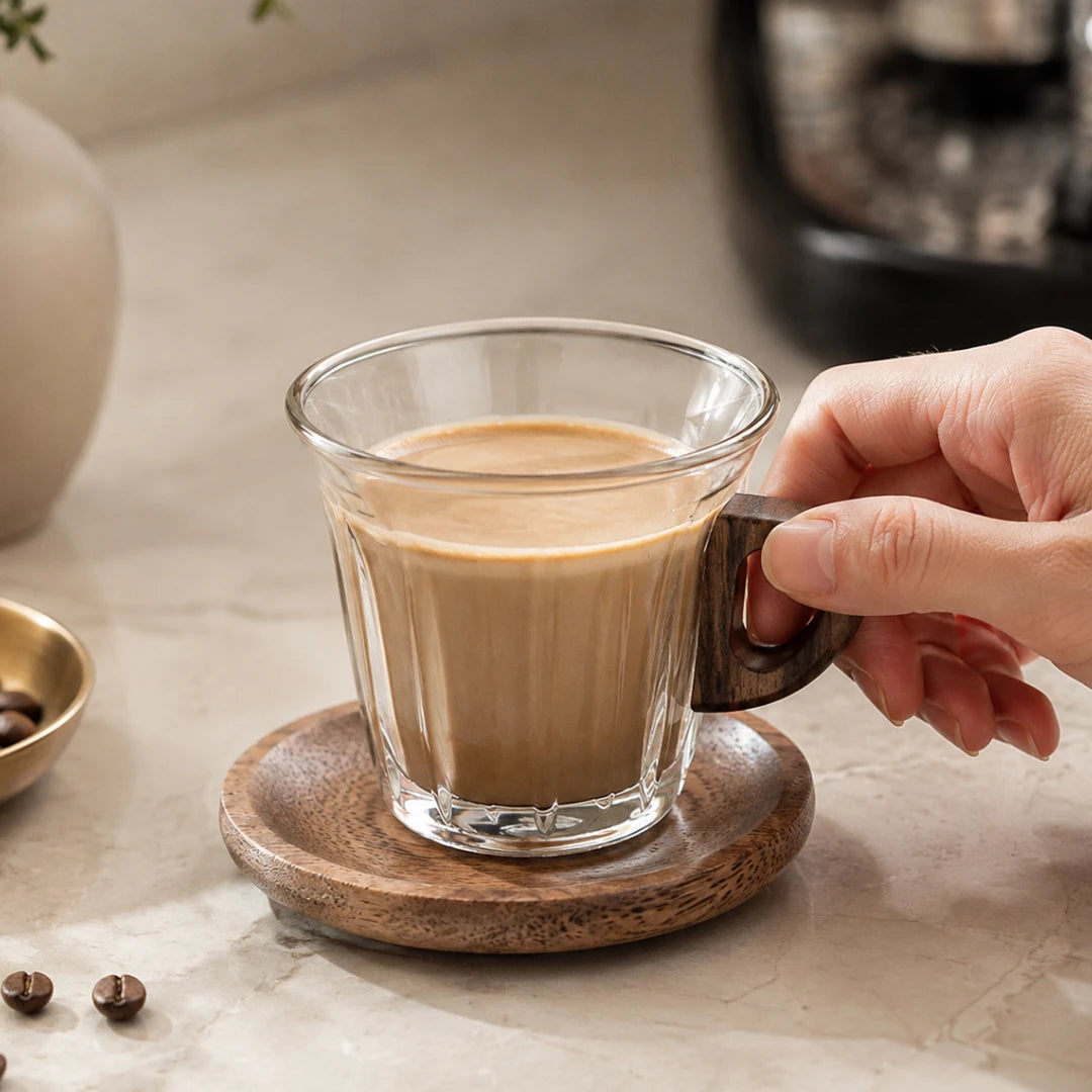 Josh & Tate - WoodGrip™ Coffee Cup Set - A hand holds the handle of a clear glass mug filled with coffee, resting on a round wooden coaster on a light stone surface.