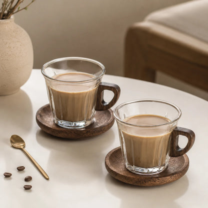 Josh & Tate - WoodGrip™ Coffee Cup Set - Two clear glass coffee mugs filled with coffee rest on round wooden coasters on a white table, with a spoon and coffee beans nearby and a softly blurred indoor setting in the background.