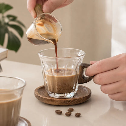 Josh & Tate - WoodGrip™ Coffee Cup Set - A person pouring dark coffee from a small glass pitcher into a clear glass mug with a dark wooden handle, placed on a round wooden coaster on a tabletop.