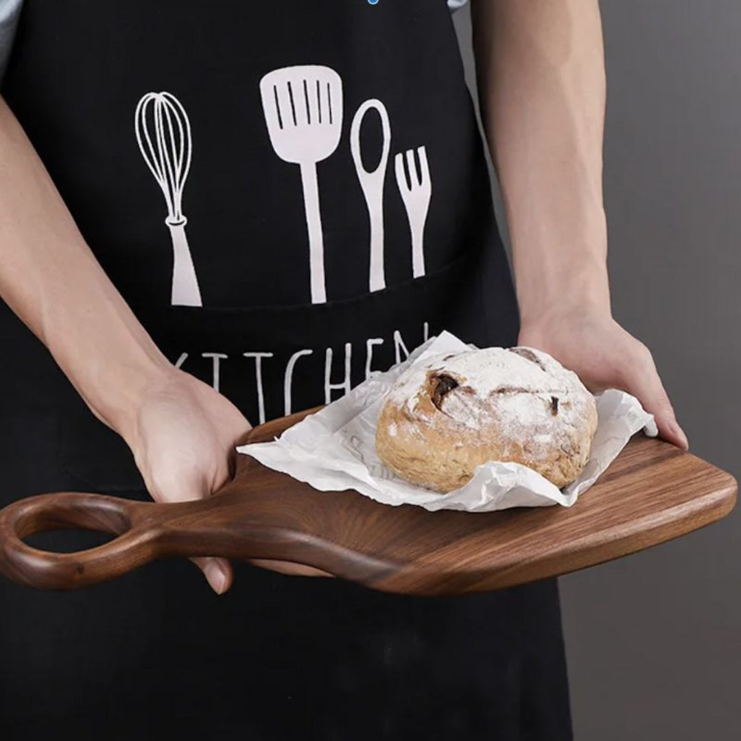Person holding a wooden board with a loaf of bread, wearing a black apron with kitchen utensil designs.