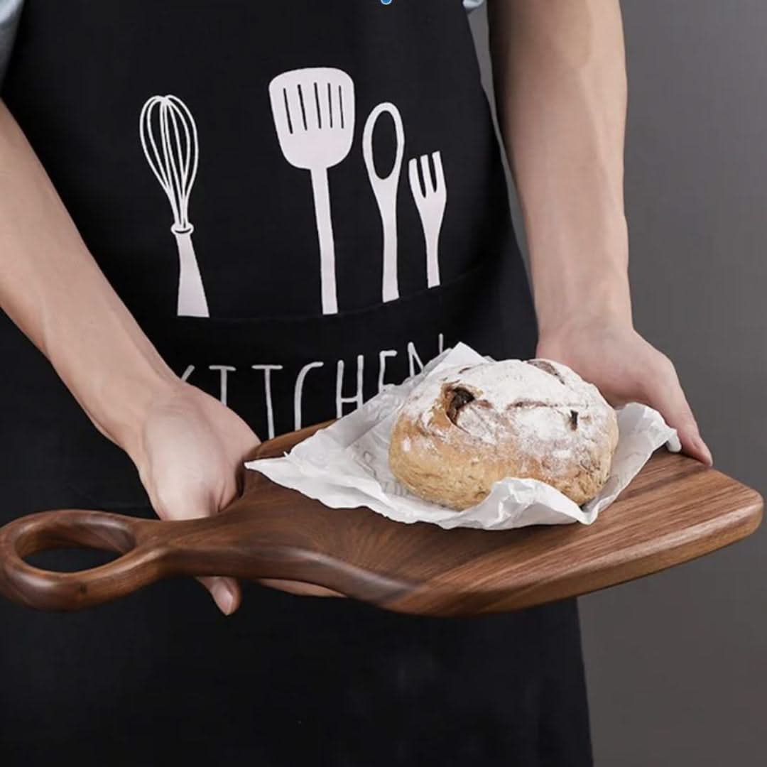Person holding a wooden board with a loaf of bread, wearing a black apron with kitchen utensil designs.