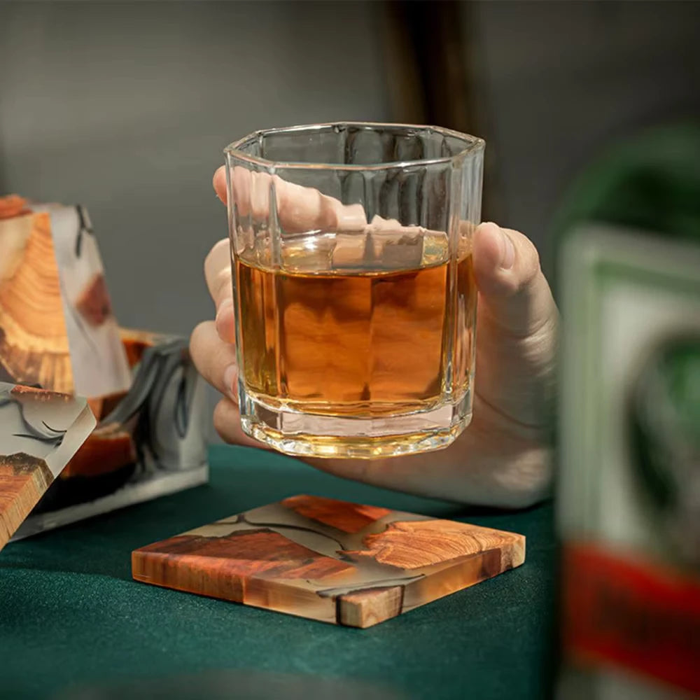 Hand holding a glass of amber liquid on a wooden coaster with a blurred background