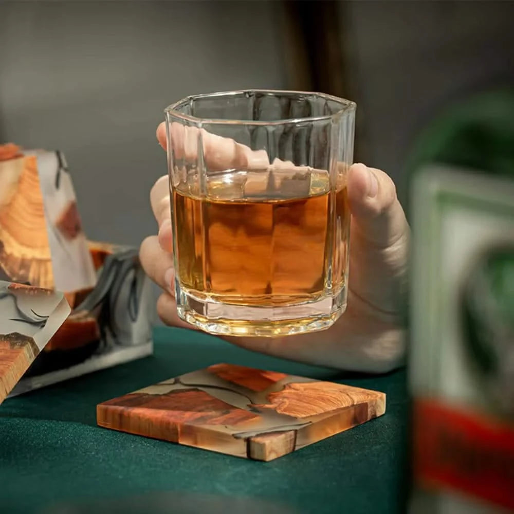 Hand holding a glass of amber liquid on a wooden coaster with a blurred background