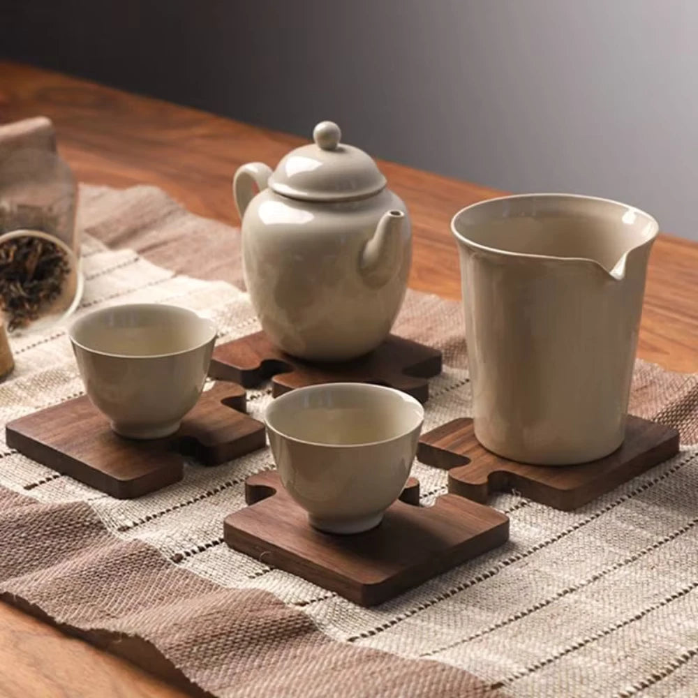 Tea set with ceramic teapot, cups, and a large cup on wooden coasters on a textured tablecloth.