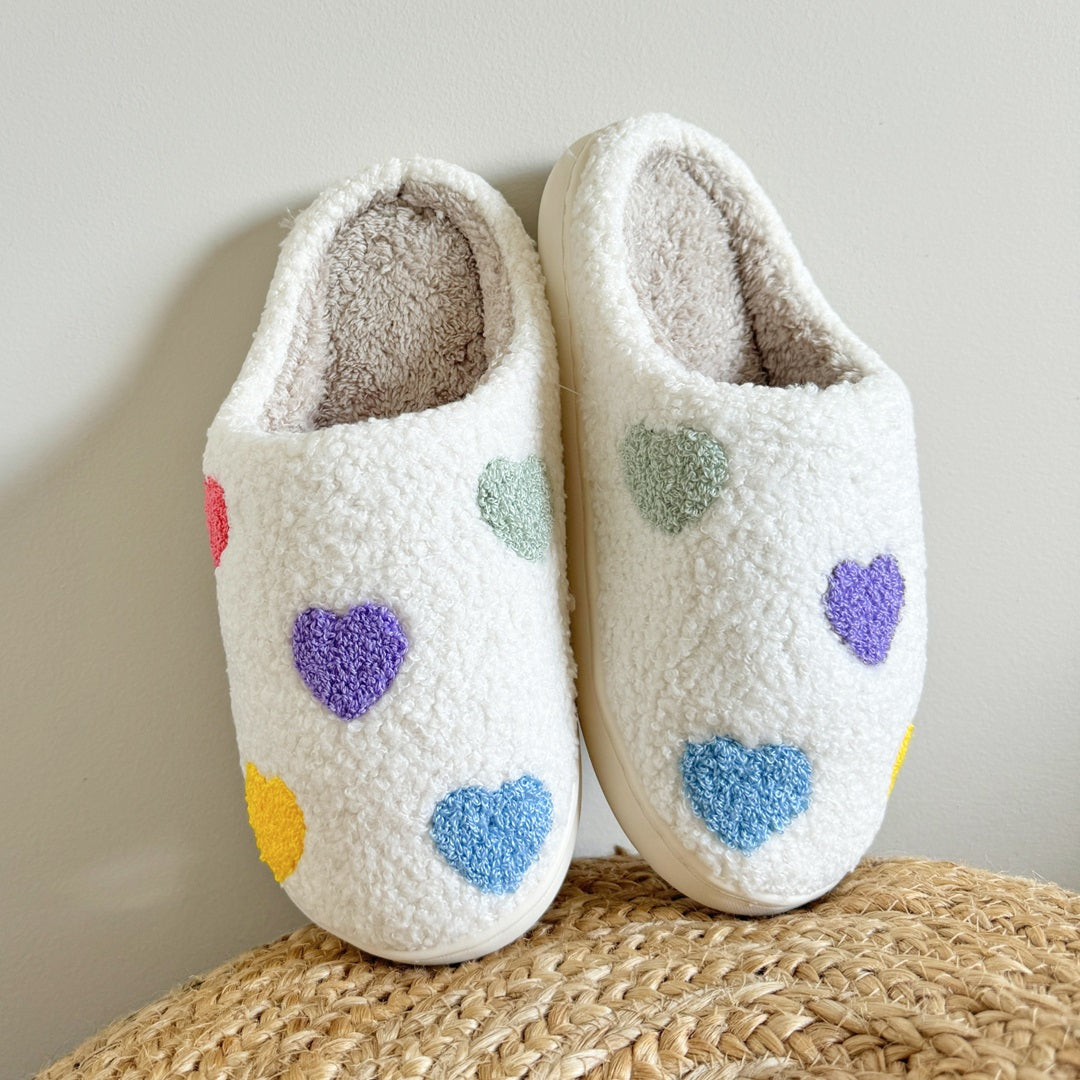 Pair of white slippers with colorful heart patterns on a woven surface.