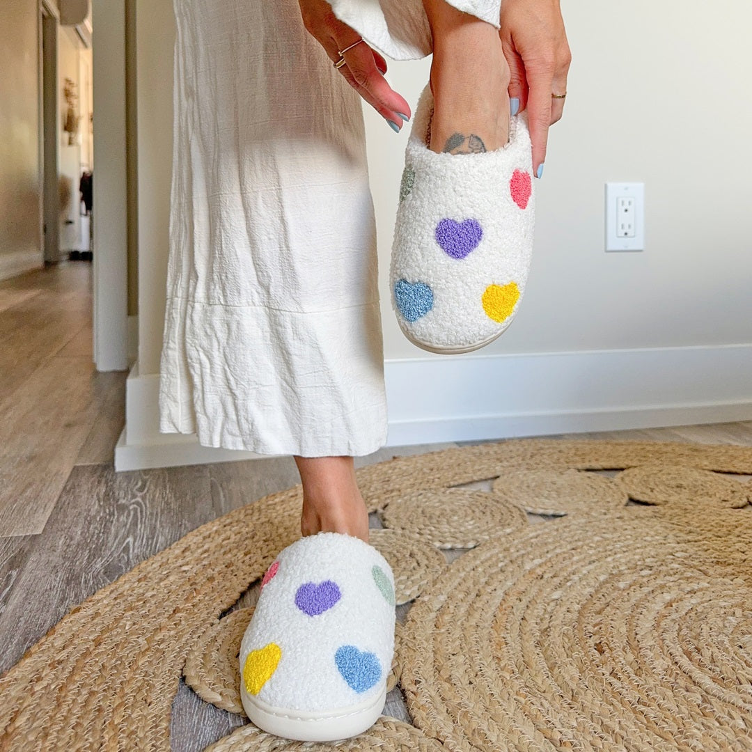 Person wearing white slippers with colorful heart patterns on a wooden floor.