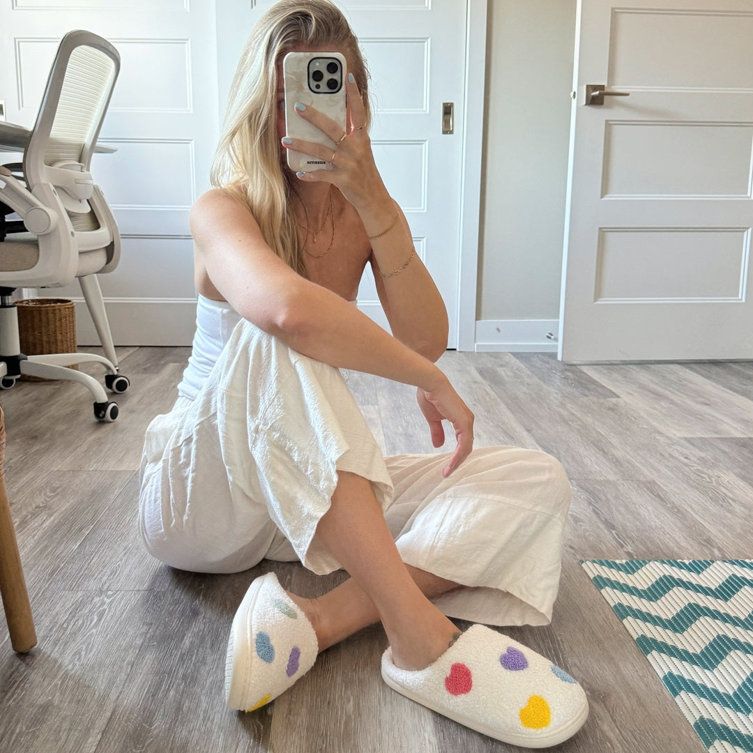 Person wearing white slippers with colorful hearts, sitting on a wooden floor.
