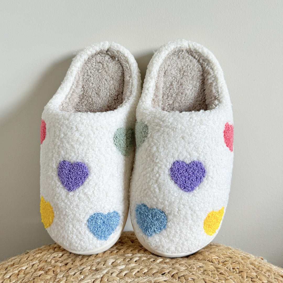 Pair of white slippers with colorful heart patterns on a woven surface.